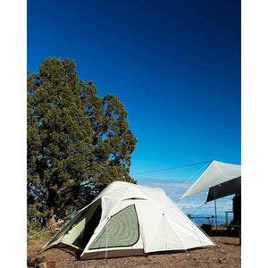 White dome tent set up on rocky ground next to a large tree under a clear blue sky