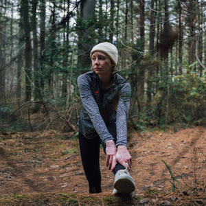 Person in a beanie and grey athletic top stretching their leg on a fallen log in a forest
