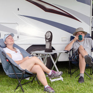 Portable desk fan on a folding table between two people sitting in camping chairs outside a recreational vehicle