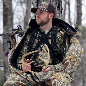 Hunter wearing Heater Body Suit camouflage gear holding deer antlers in a tree stand with a compound bow