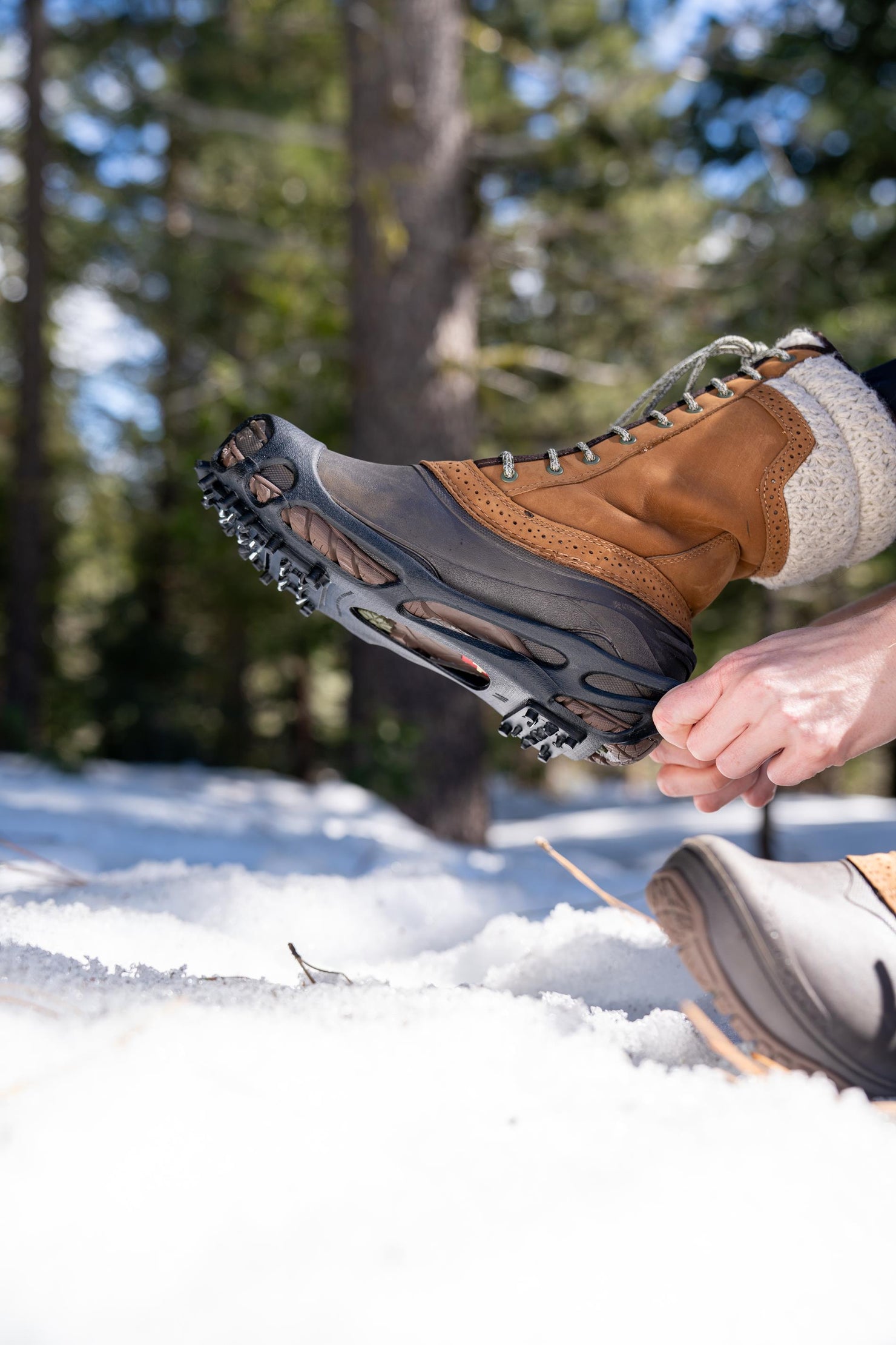 Person attaching black ice cleats to a brown winter boot in the snow
