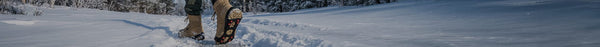 A person wearing tan boots with ice cleats walks through deep, sunlit snow, leaving footprints in a winter forest.