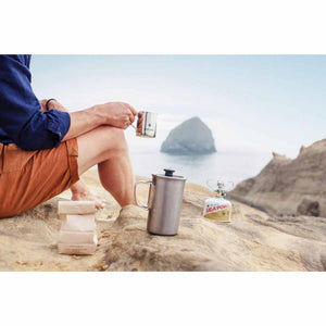 Person holding a Snow Peak mug near a GigaPower stove and coffee press on a rocky beach