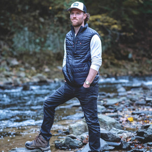 Man wearing a black puffer vest white long sleeve shirt and The North Face cap standing by a rocky stream