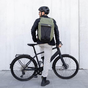 Person wearing a green Thule backpack and black helmet standing with a black bicycle