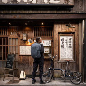 Person wearing a Thule backpack standing next to a folding bicycle in front of a wooden storefront