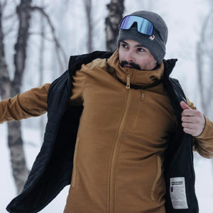 Person wearing a Fjallraven brown fleece jacket and black vest with a grey beanie and ski goggles in a snowy forest