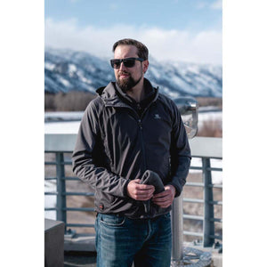 Man wearing a grey hooded jacket and sunglasses standing outdoors with snowy mountains in the background