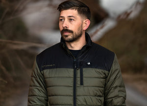 A man with a beard and dark hair wears an olive green and black puffer jacket with "ACTIONHEAT™" text, looking left against a blurred outdoor background.