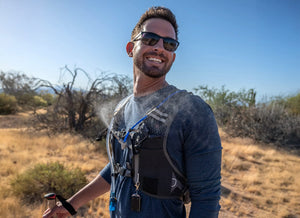 A smiling man in sunglasses and a misting hydration vest sprays mist while hiking in a dry, sunny landscape.