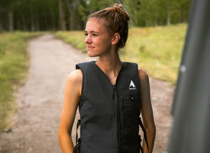 A young woman with a messy bun smiles, looking left, wearing a black Alphacool cooling vest on a dirt path with blurred trees.