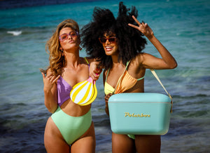 Two women in colorful swimsuits pose playfully on a beach, one holding a beach paddle and the other a light blue "Polarbox" cooler, with blue ocean behind them.