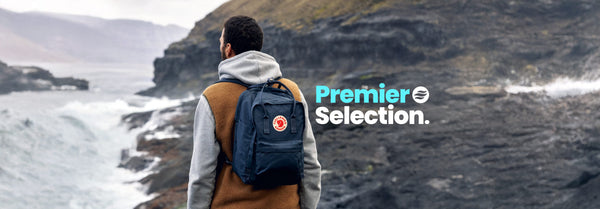 From behind, a man in a grey hoodie and brown vest with a blue backpack views a stormy, rocky coastline. Text: 