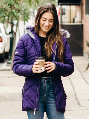 A smiling young woman in a purple puffer jacket with a fur hood holds a coffee cup outdoors on a sidewalk.