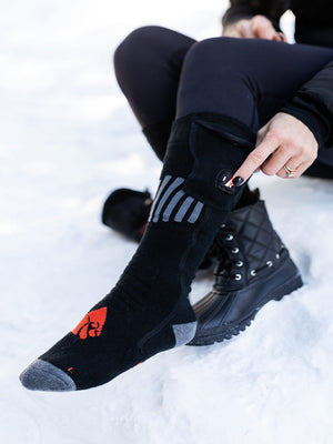 A person sits in snow, pressing the glowing flame button on a black heated sock with gray stripes and a red logo, worn with black quilted boots.