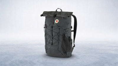 A dark grey Fjällräven Atasko 25 rolltop backpack with a bungee cord system and mesh pockets stands on a light grey textured surface.