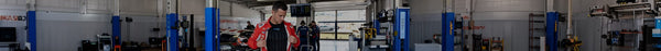 A man in a red racing jacket stands in a professional race car garage with a car, crew, and equipment. A banner reads 