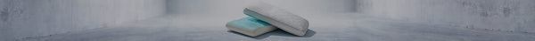Two stacked rectangular pillows, the bottom one white with a light blue top, the top one light gray with a chevron pattern, on a grey floor.