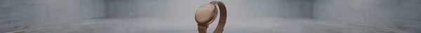 A rose gold smartwatch with a mesh band stands upright in a clean, grey minimalist setting.