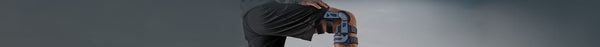A person's bent leg in black shorts wears a black and grey knee brace, with a hand resting on their upper thigh against a grey background.
