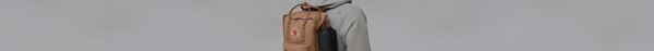 A person in a light grey hoodie wears a tan Fjällräven Kånken backpack, with a black water bottle in its side pocket, against a plain grey background.