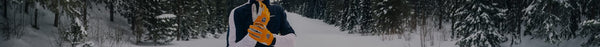 A person in a snowy forest, wearing a dark blue jacket and bright orange gloves with 