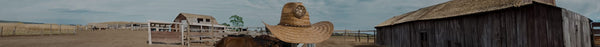 Panoramic view of a rustic ranch with a straw cowboy hat, a horse, wooden fences, and weathered barns under a cloudy sky.