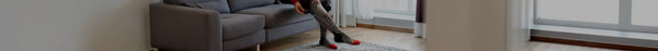 A person adjusts gray compression socks with red accents and black straps, wearing red toe socks, while seated beside a gray sofa.