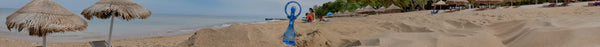 Panoramic view of a sandy beach with straw umbrellas, a blue beach toy, people, and lounge chairs, with the ocean and green trees in the background.