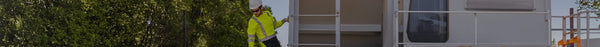 A worker in a yellow safety shirt and hard hat climbs or inspects a white modular building with railings, lush green trees in the background.