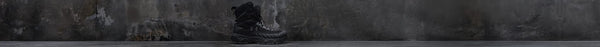 A single black tactical boot stands on a dark concrete floor against a distressed dark grey concrete wall, with the Under Armour logo visible.