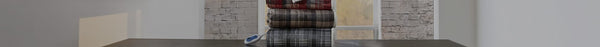 A stack of three plaid electric blankets (red, brown, grey) with a white remote on a dark table, in a room with a window and a light brick wall.