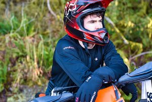 Person wearing ActionHeat black long sleeve shirt and gloves with a red motocross helmet leaning on a motorcycle