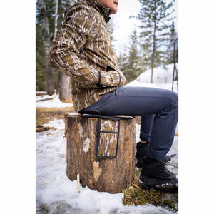 Person wearing Mobile Warming camouflage jacket sitting on a tree stump with a matching camouflage seat cushion