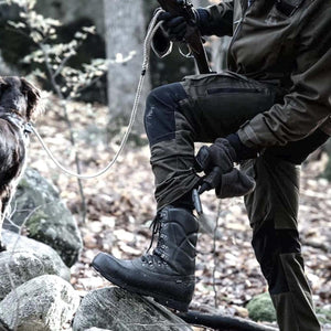 Arctic Volt hunter adjusting black leather boot while holding a rifle and dog leash in a forest