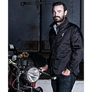 Man with beard wearing a black windbreaker jacket standing next to a motorcycle