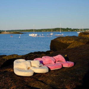 Acorn white and pink plush flip flops on a rocky shore with sailboats in the background