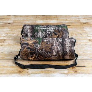 Camo hand warmer muff with Saskatchewan Original branding and black adjustable strap on wooden surface