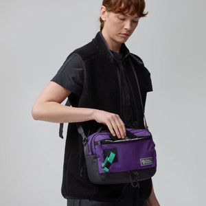 Person wearing a black fleece vest and carrying a purple Fjallraven shoulder bag