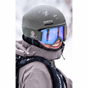 Person wearing a matte olive Smith ski helmet and blue mirrored goggles in snowy conditions