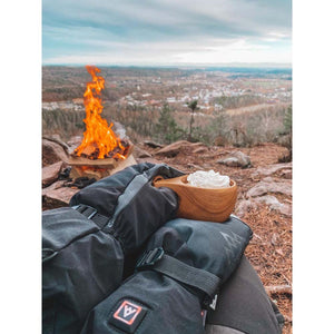 Person wearing Heat Experience black heated mittens holding a wooden cup with whipped cream by a campfire