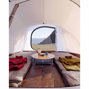 Interior view of a tent with two sleeping pads, a round wooden table, and a view of the coast through the open door