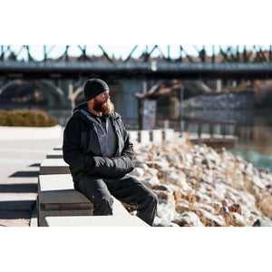 Man with beard wearing black beanie and jacket sitting on concrete wall by river
