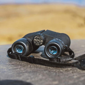 Black NOCS Provisions binoculars with strap resting on a stone surface