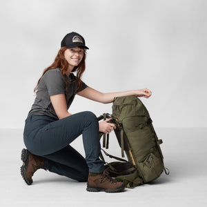 Woman kneeling beside a green Fjallraven hiking backpack wearing a cap and outdoor gear