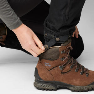 Hand adjusting the snap button strap on the hem of black FjallRaven trousers over a brown Hanwag hiking boot