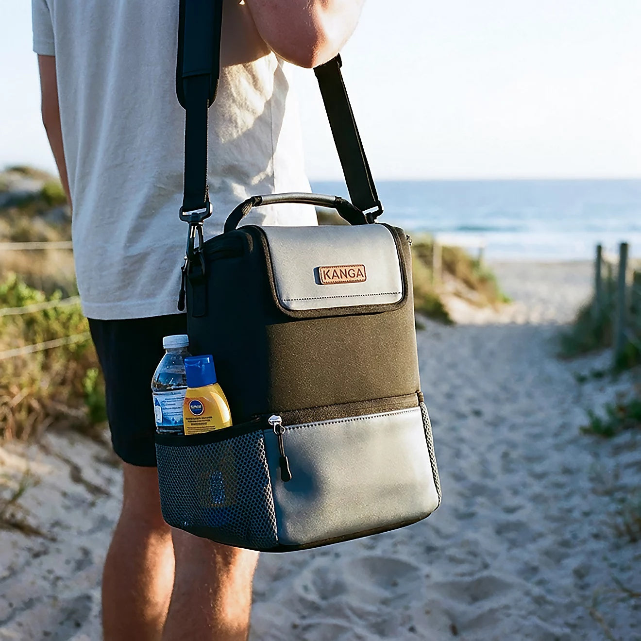 Person carrying a Kanga cooler bag with a water bottle and sunscreen in the side mesh pocket on a sandy beach path
