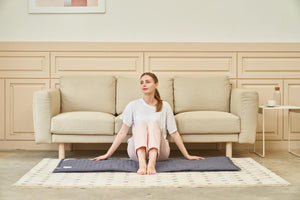 Woman sitting on a grey quilted mat on a rug in front of a beige sofa