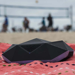 Black geometric portable speaker on a red beach towel with a blurred volleyball net in the background