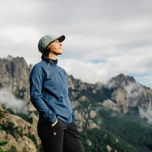 Person wearing a blue Fjallraven fleece jacket and cap standing in front of mountain peaks